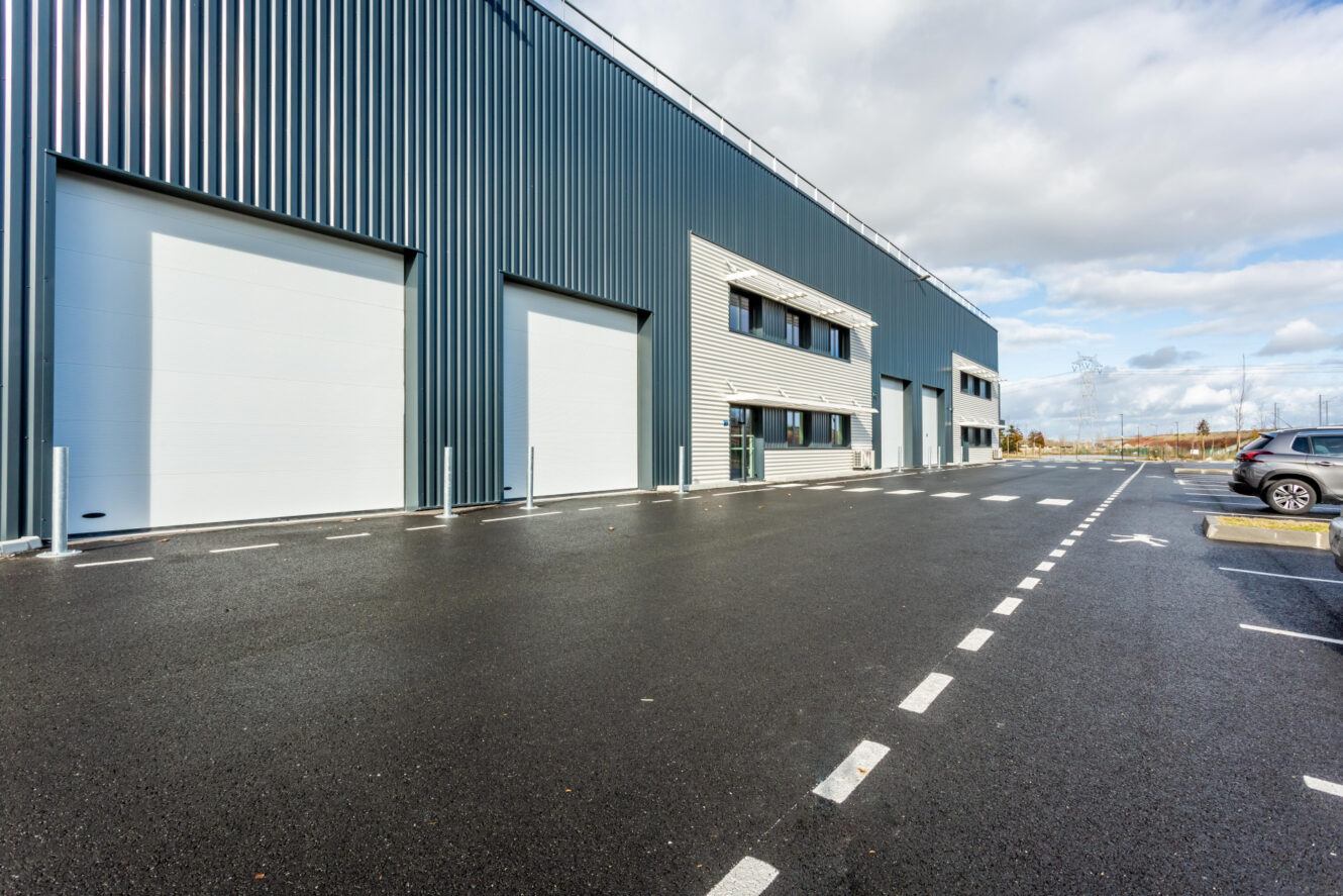 Vue extérieure d'un entrepôt industriel moderne avec de grandes portes de garage et un parking pavé vide sous un ciel partiellement nuageux.
