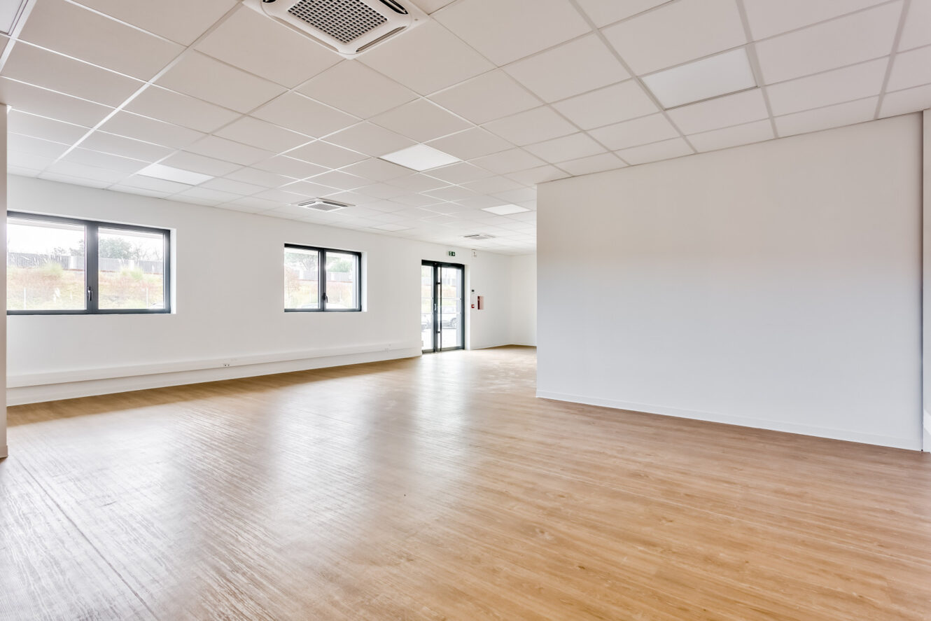 Pièce vide et moderne avec de grandes fenêtres, un parquet en bois clair, des murs blancs et un plafond suspendu avec des éclairages encastrés.