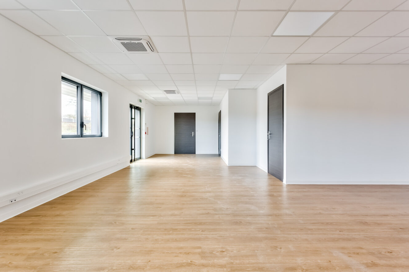 Pièce vide et moderne avec un sol en bois clair, des murs blancs, plusieurs portes sombres, des panneaux au plafond et de grandes fenêtres sur le côté gauche pour laisser passer la lumière naturelle.