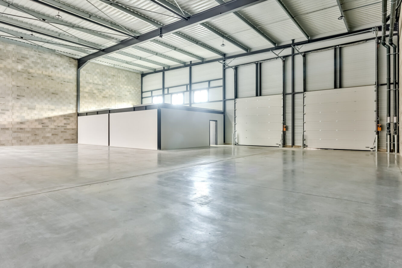 Entrepôt industriel vide avec sol en béton, hauts plafonds, grandes portes de garage, et un espace de bureau fermé avec des fenêtres et une porte à l'intérieur de l'entrepôt.