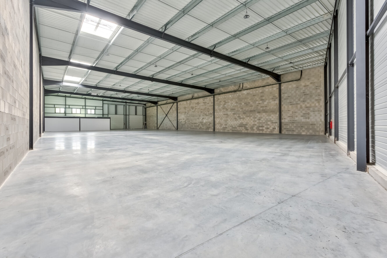 Entrepôt industriel vide avec des sols en béton, de hauts plafonds, des poutres métalliques apparentes et de grandes portes roulantes le long d'un mur. Les murs sont faits de blocs de béton. L'espace est bien éclairé.