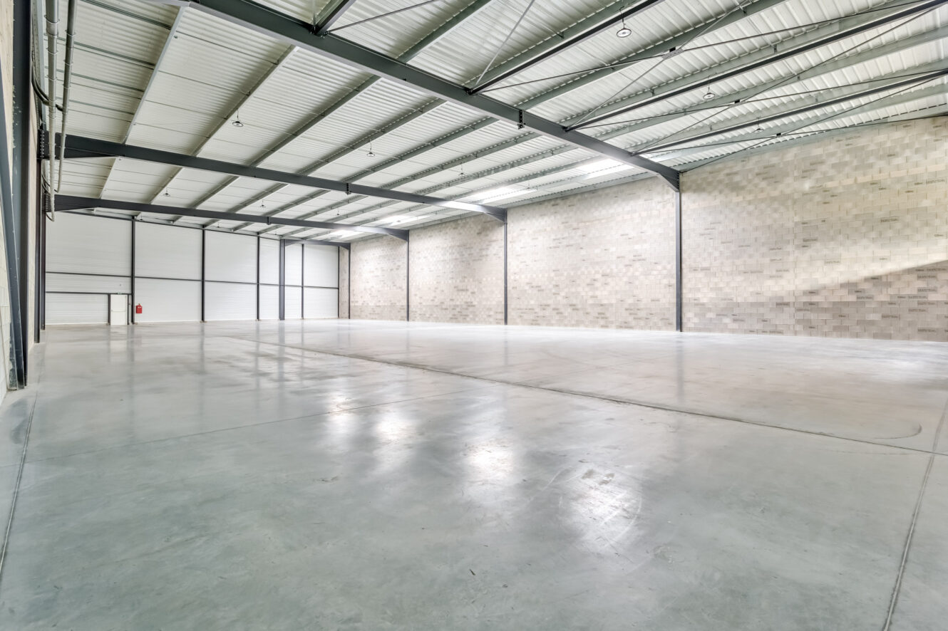 Entrepôt industriel vide avec sol en béton poli, haut plafond métallique et murs en briques. De grandes portes coulissantes et un extincteur sont visibles sur la gauche.