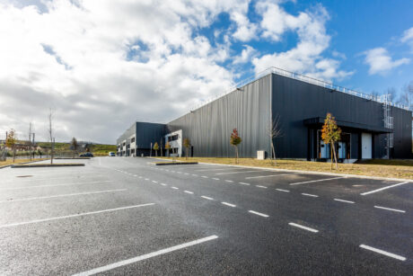 Un grand bâtiment industriel moderne à l'extérieur gris et un parking vide sous un ciel partiellement nuageux.