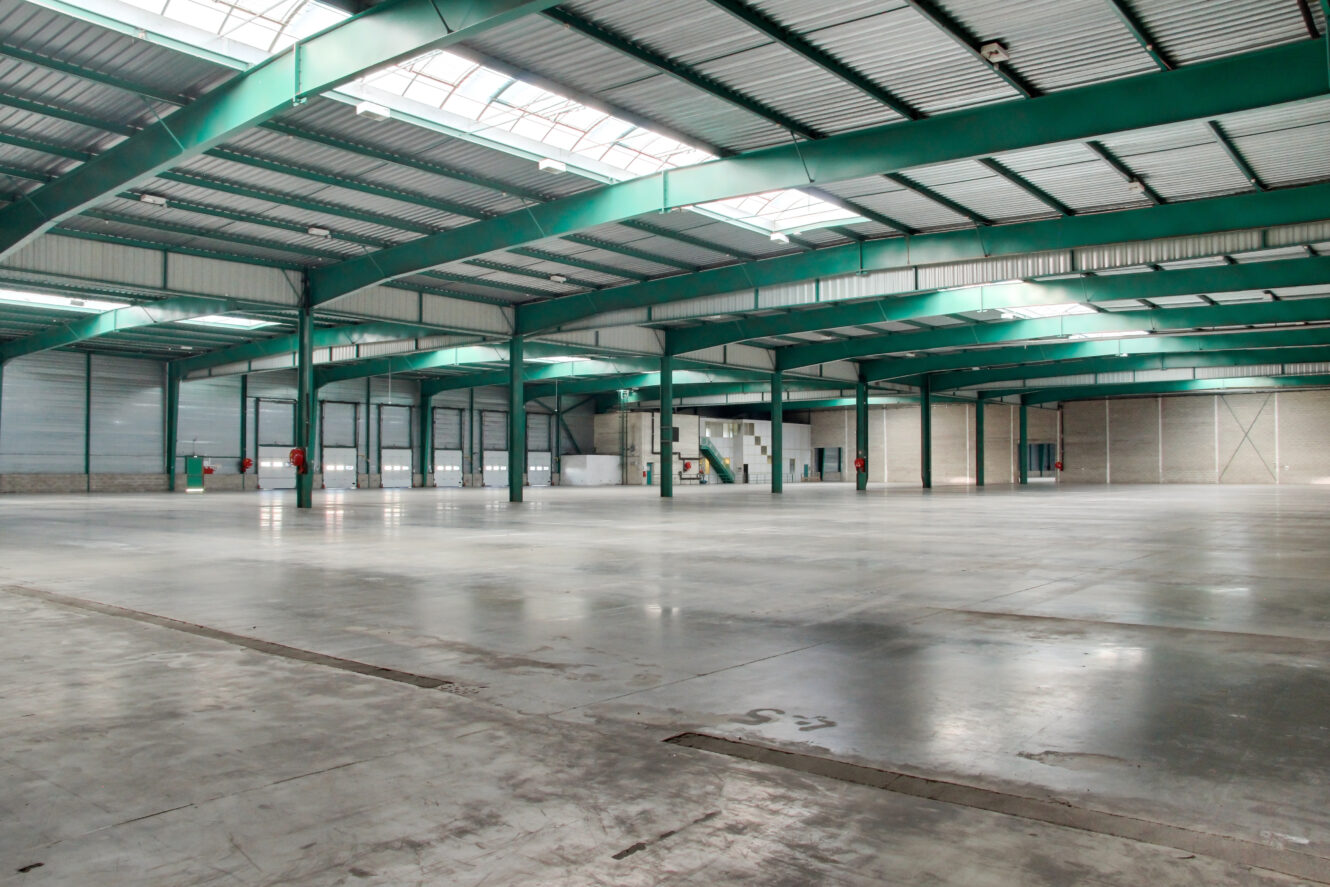 Grand entrepôt industriel vide avec des poutres métalliques vertes, de hauts plafonds, un sol en béton et des puits de lumière permettant un éclairage naturel à l'intérieur.