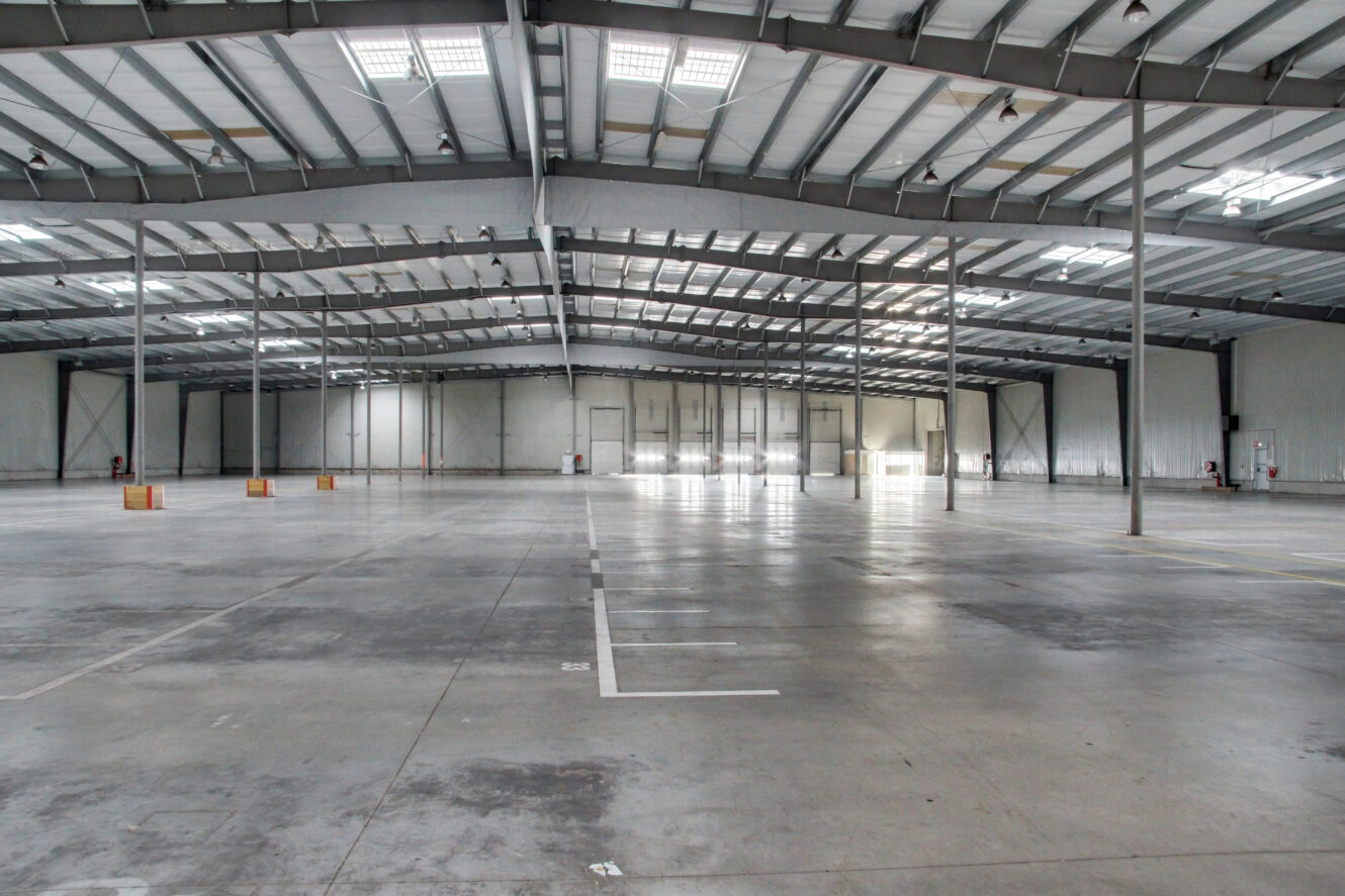 Intérieur d'un grand entrepôt vide avec de hauts plafonds, des poutres apparentes, un sol en béton, des colonnes de soutien et des éclairages aériens ; aucun équipement ou inventaire n'est présent.