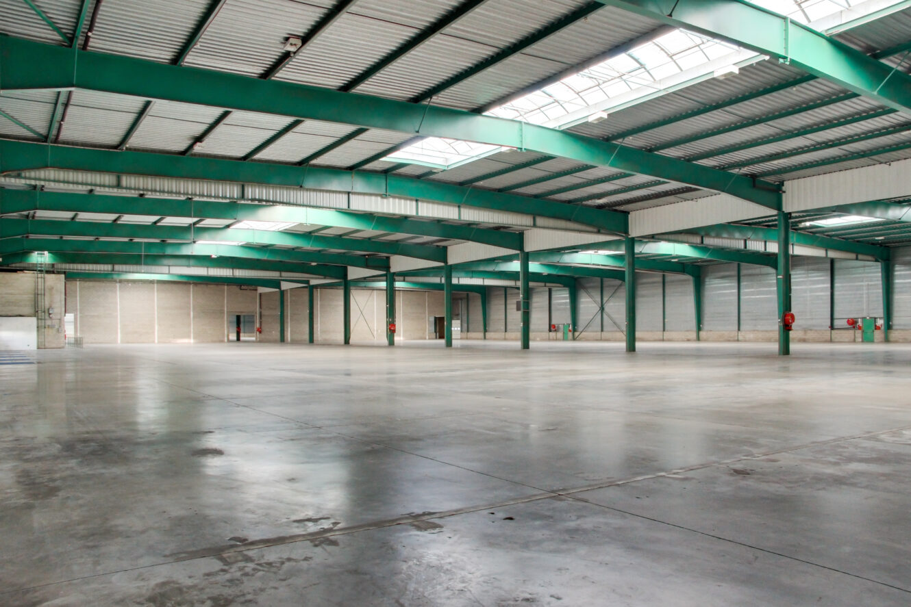 Intérieur d'un grand entrepôt vide avec des poutres métalliques vertes, un sol en béton, un haut plafond et des lucarnes laissant entrer la lumière naturelle.