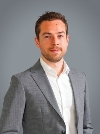 Un homme portant une veste de costume gris clair et une chemise blanche se tient sur un fond gris uni, regardant l'appareil photo avec une expression neutre.