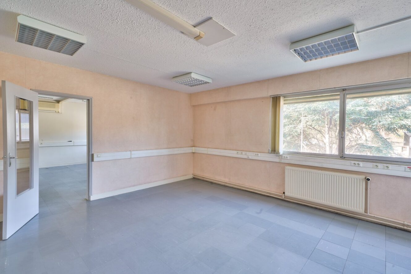 Une pièce vide aux murs clairs, au sol carrelé gris, aux plafonniers fluorescents, un radiateur sous une grande fenêtre et une porte ouverte menant à un autre espace vide.