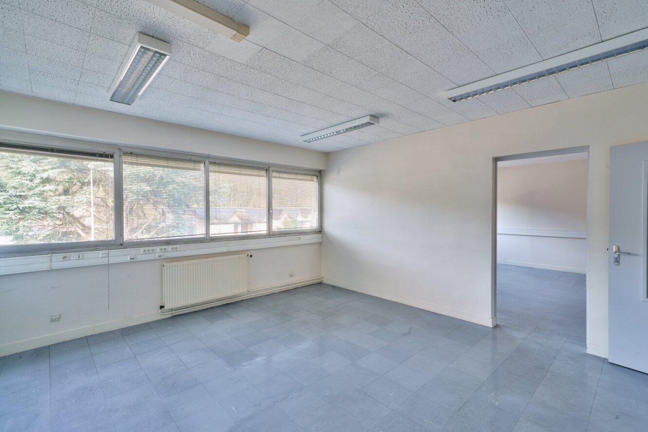 Bureau vide avec sol carrelé, radiateur, plafonniers fluorescents, grandes fenêtres avec stores, et une porte ouverte menant à une autre pièce.