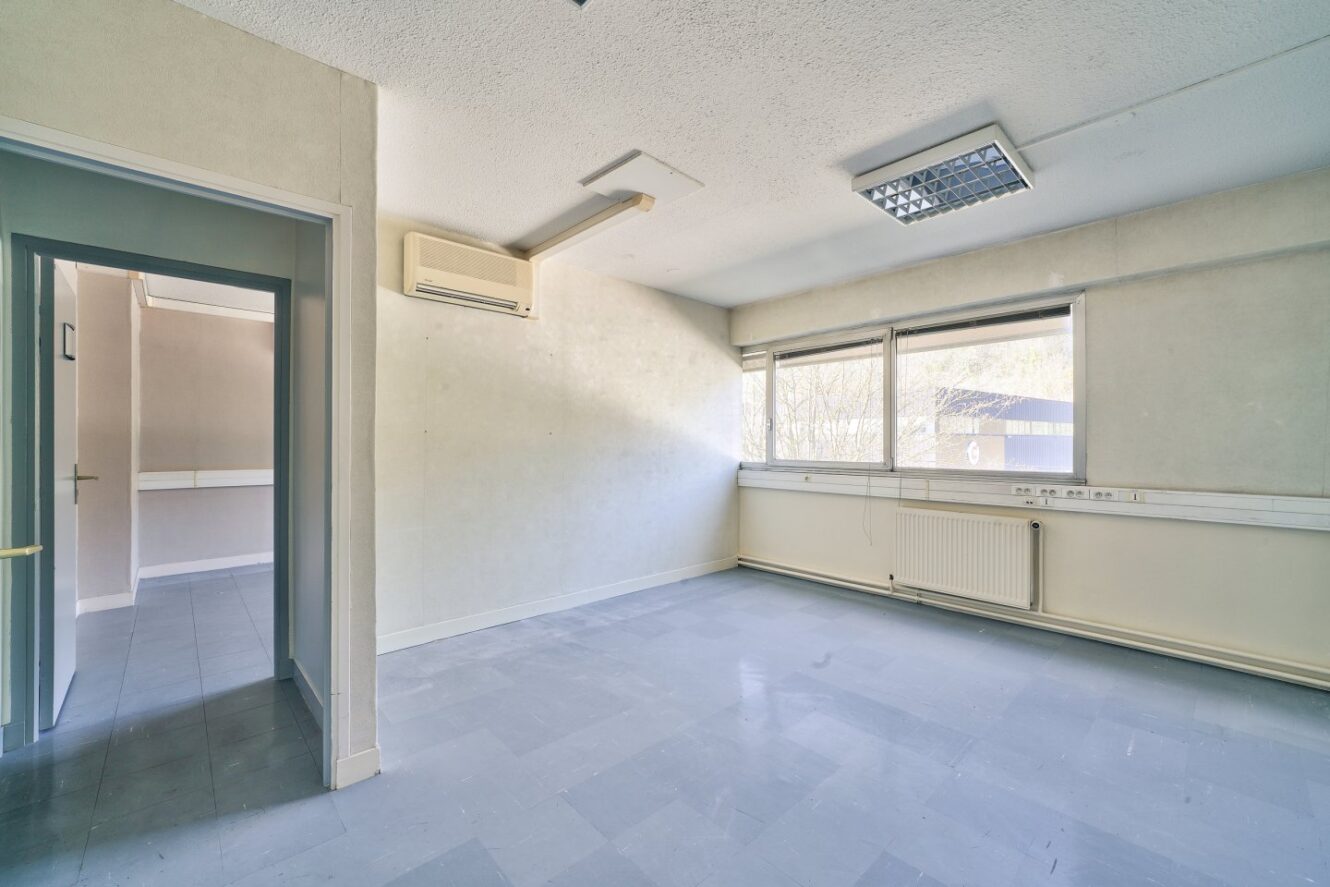 Pièce vide, non meublée, avec un sol en carrelage gris, des murs blancs, deux grandes fenêtres, un radiateur et un climatiseur mural. Une porte ouverte mène à un couloir.