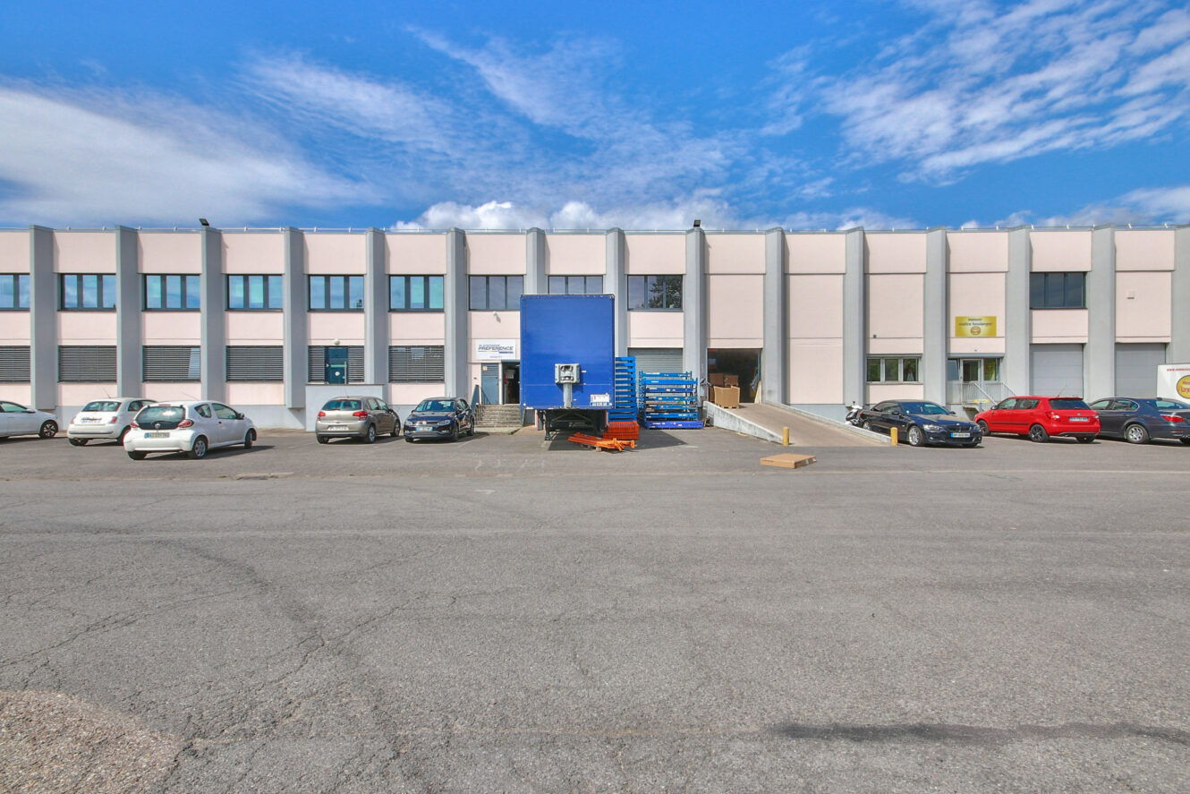 Un grand entrepôt avec de nombreuses fenêtres, des voitures garées devant et un quai de chargement avec des palettes bleues empilées.