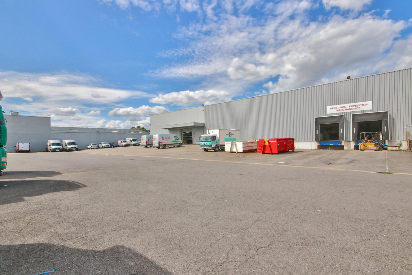 Grand entrepôt industriel avec un revêtement gris, des bennes à ordures rouges, plusieurs camions et camionnettes garés sous un ciel partiellement nuageux. Le panneau au-dessus de la porte indique "Réception - Expédition".