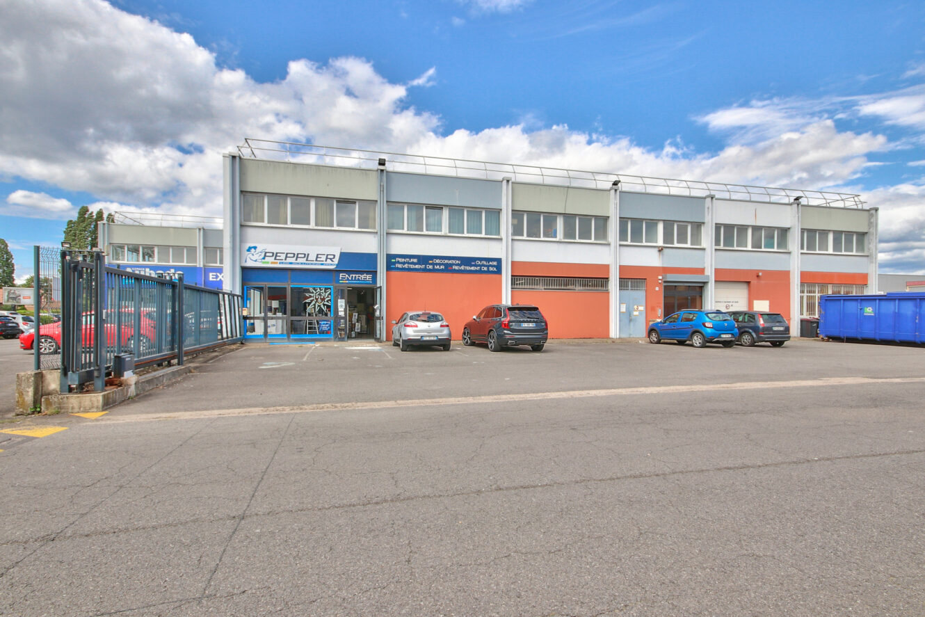 Un bâtiment commercial avec de grandes fenêtres, plusieurs voitures garées, une benne à ordures bleue et un panneau indiquant "Peppler" sur la façade, sous un ciel partiellement nuageux.