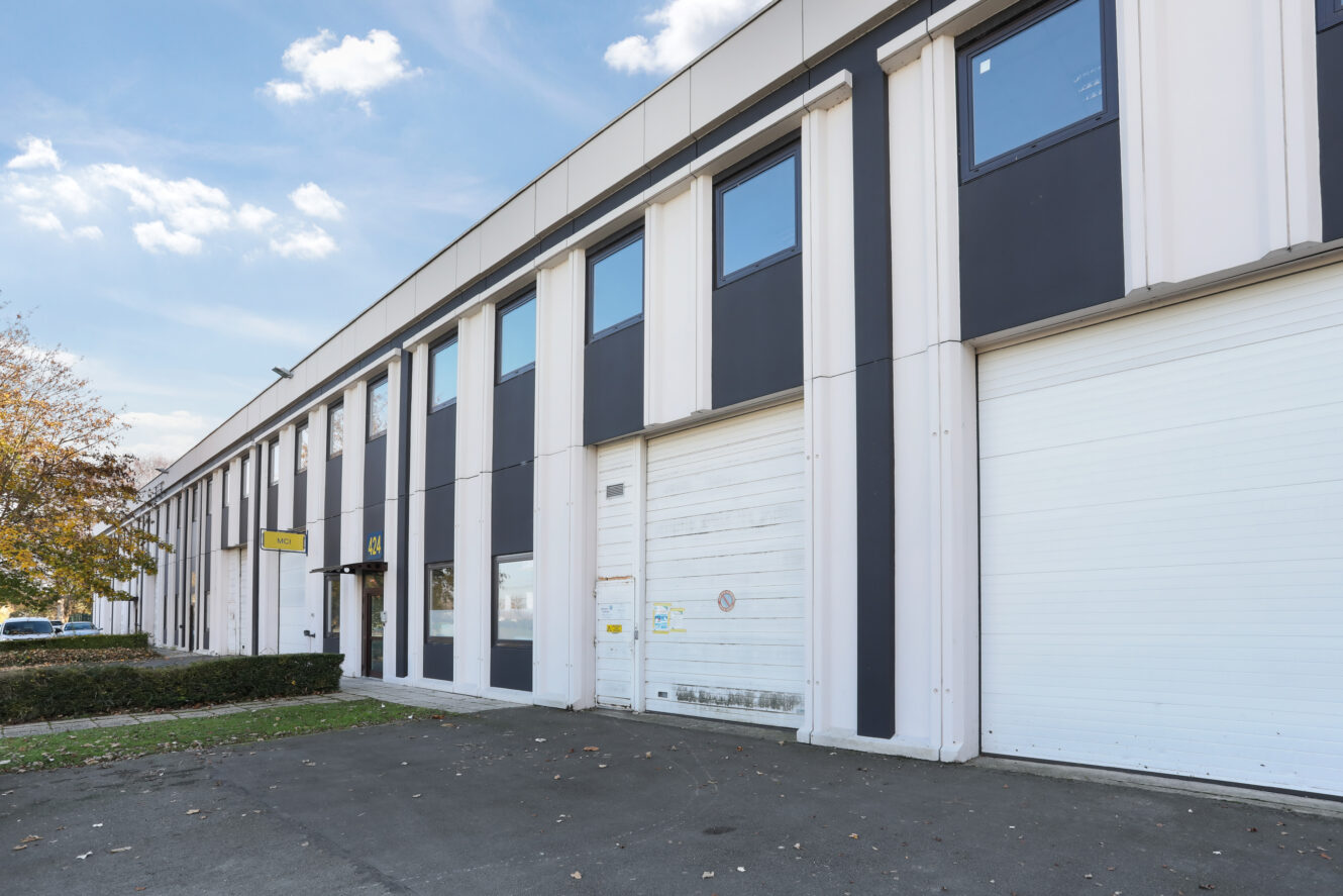 Un entrepôt industriel moderne avec de grandes portes de garage blanches et des fenêtres supérieures, situé à côté d'un terrain pavé et d'une petite haie.