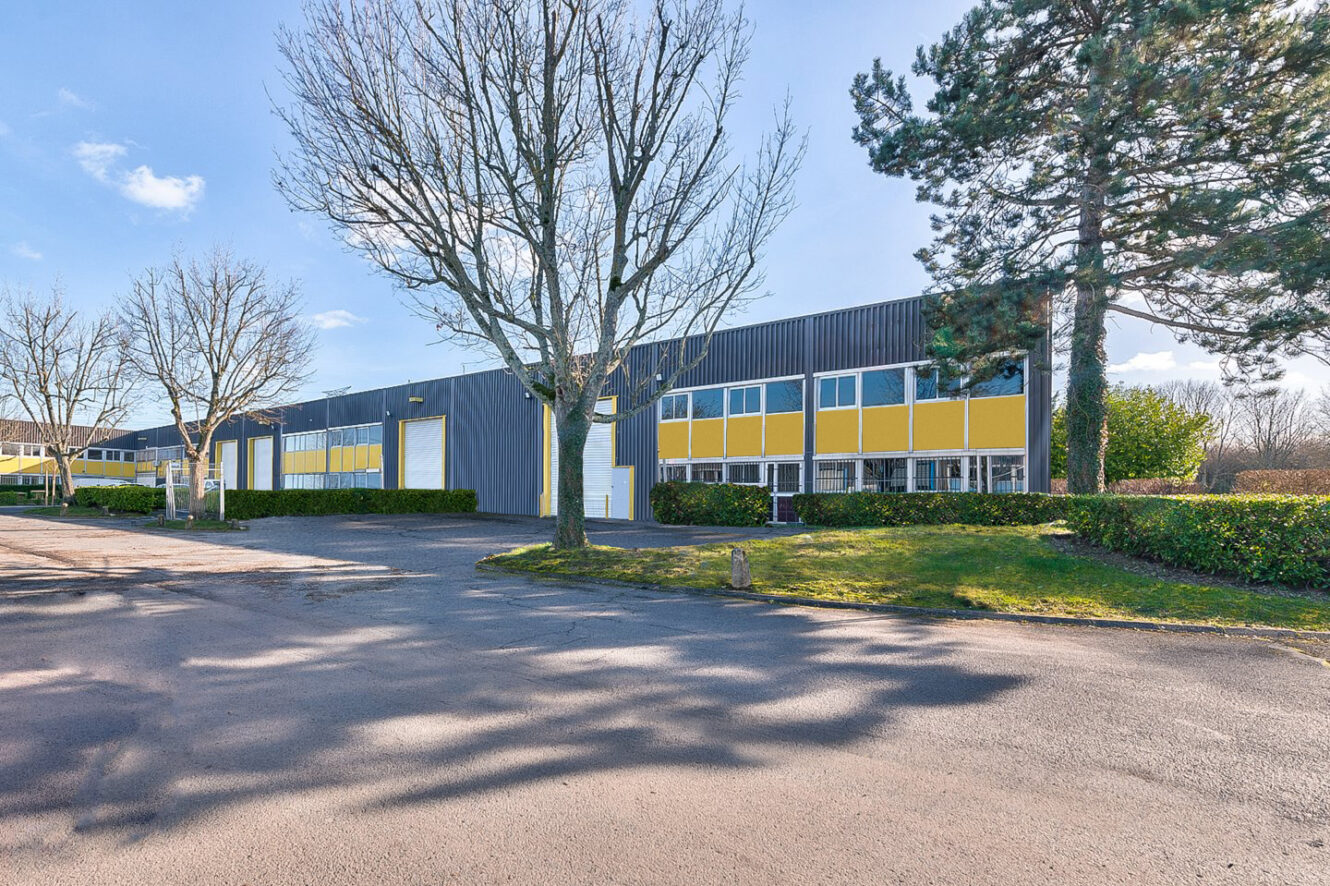 Un long bâtiment industriel avec des panneaux jaunes et gris, entouré d'arbres et d'un parking pavé sous un ciel clair.