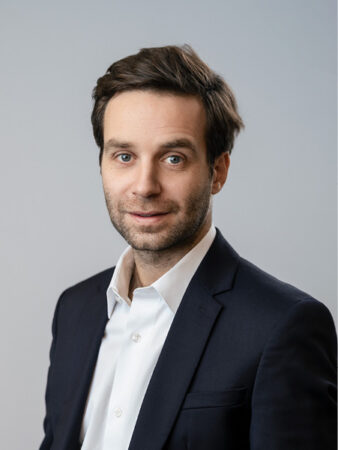 Un homme aux cheveux bruns courts et à la barbe, vêtu d'une veste de costume sombre et d'une chemise blanche, pose sur un fond clair uni.