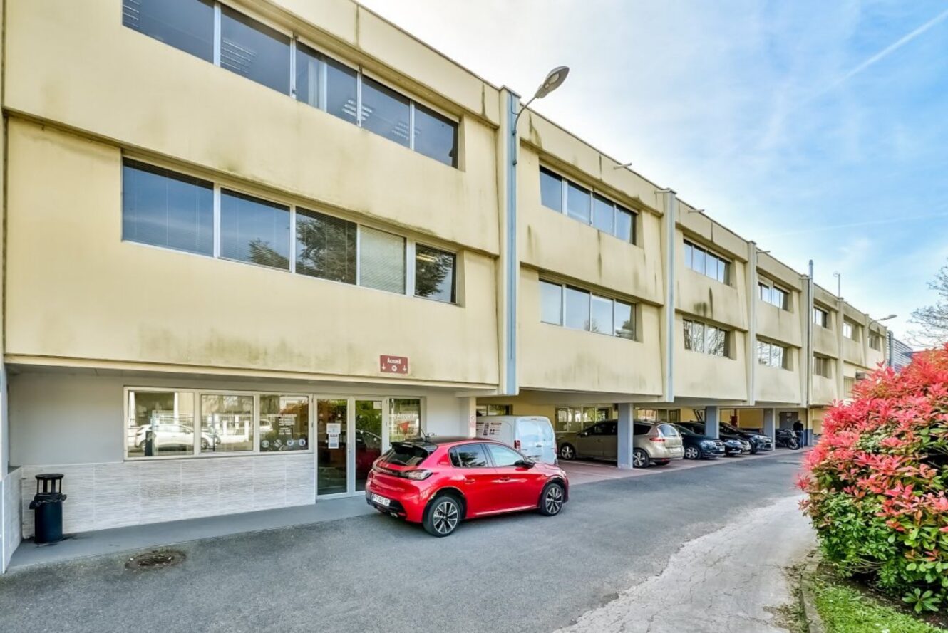 Un bâtiment beige de deux étages avec de grandes fenêtres, un parking au rez-de-chaussée, plusieurs voitures garées et une voiture rouge à l'avant. Des buissons et un lampadaire sont visibles sur le côté droit.