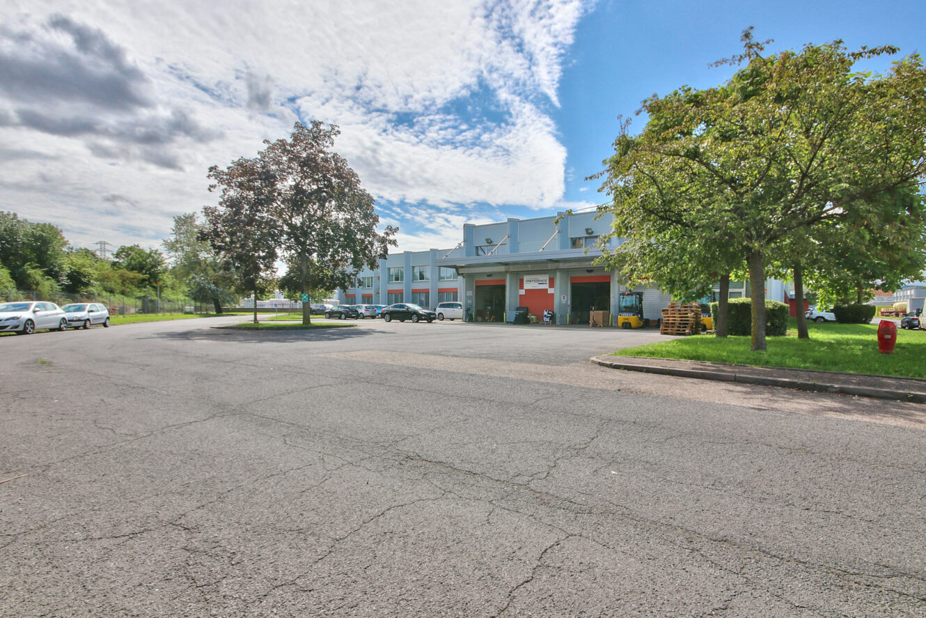 Grand parking avec des voitures éparpillées devant un bâtiment commercial sous un ciel bleu avec des nuages et plusieurs arbres le long de la chaussée.