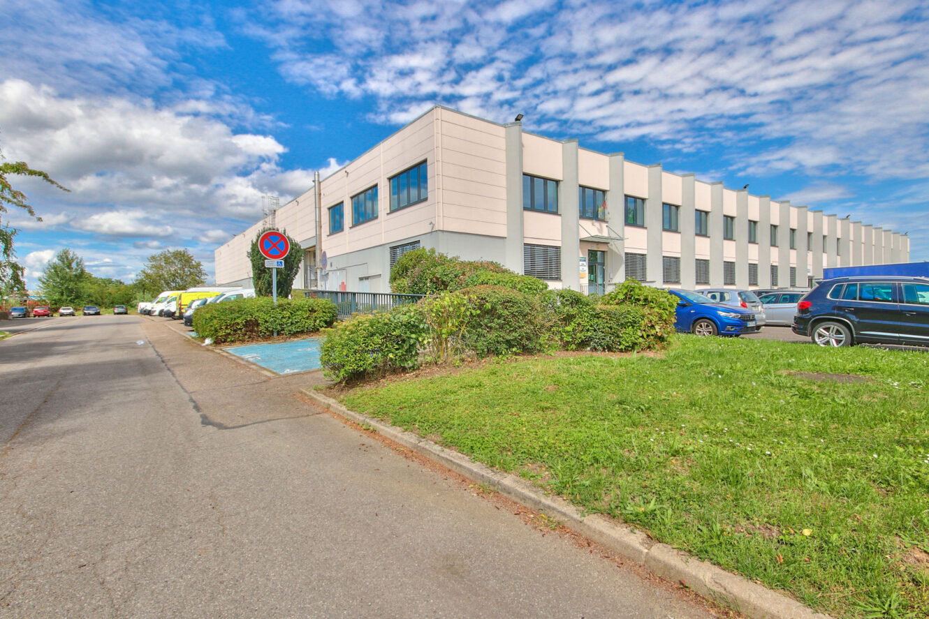 Un bâtiment commercial moderne de deux étages avec de grandes fenêtres, bordé d'une pelouse verte et de voitures garées, sous un ciel partiellement nuageux.