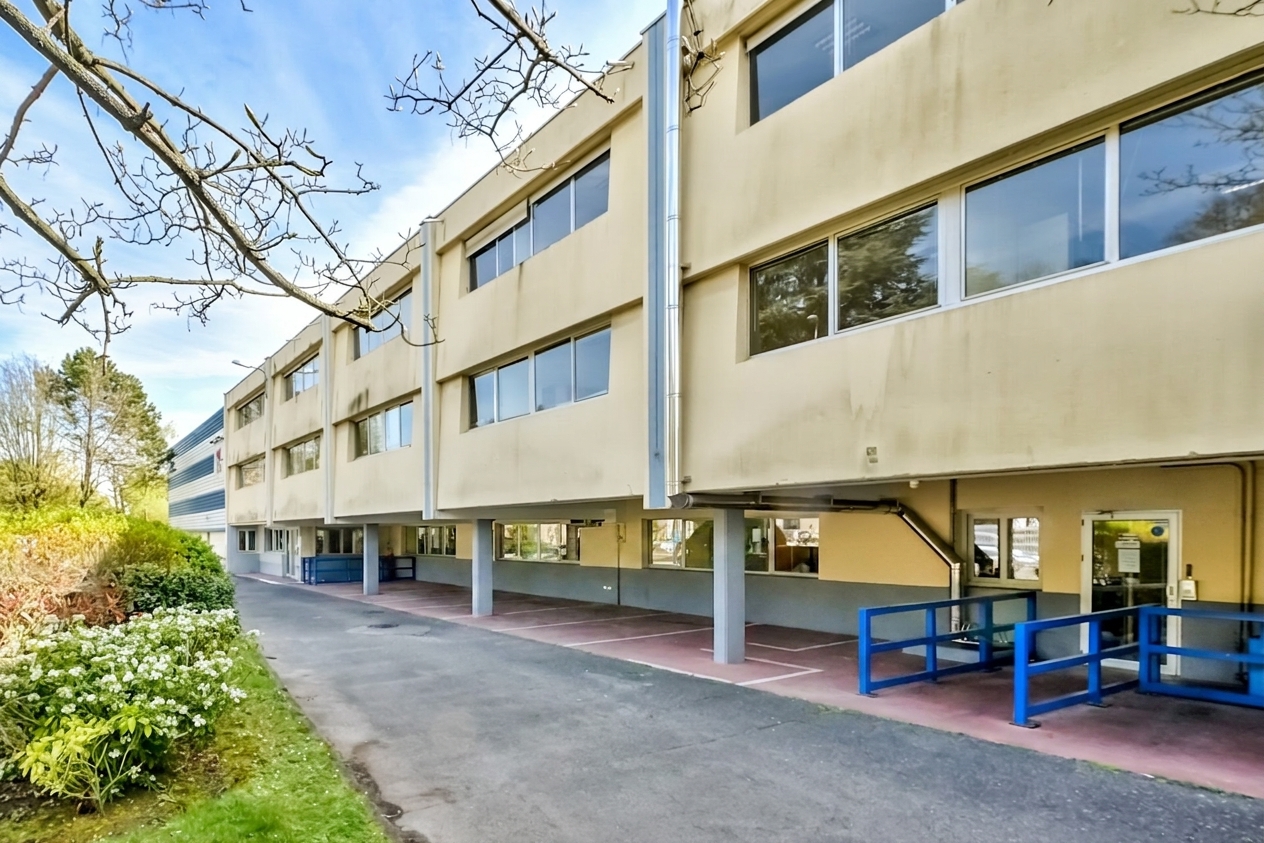 Un bâtiment beige de deux étages, doté de grandes fenêtres et reposant sur des piliers, se dresse à côté d'une allée pavée et d'un espace vert aménagé, sous un ciel dégagé.