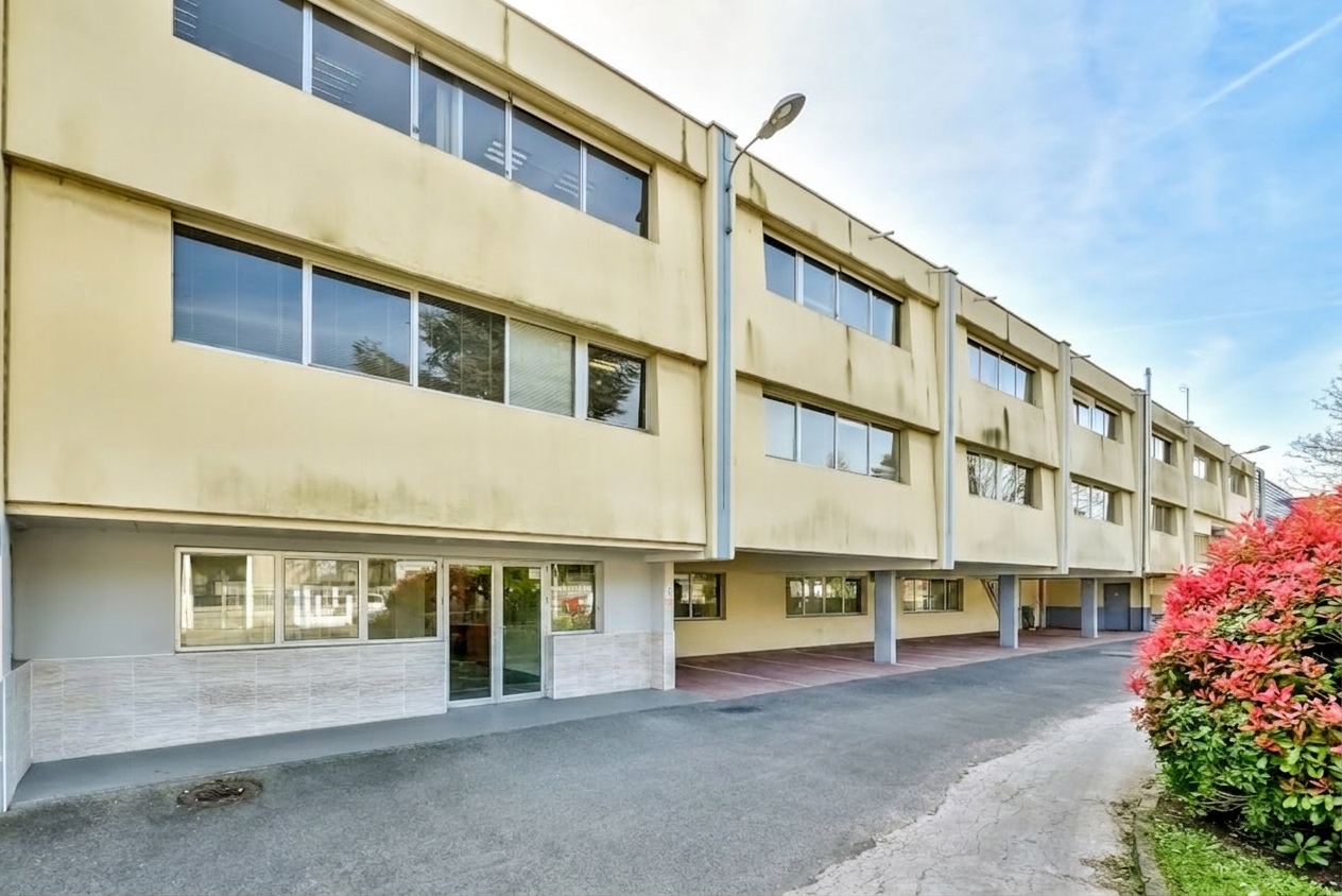 Un immeuble jaune de trois étages, doté de grandes fenêtres et d'un parking couvert au rez-de-chaussée, situé à côté d'une allée pavée et bordé de buissons.