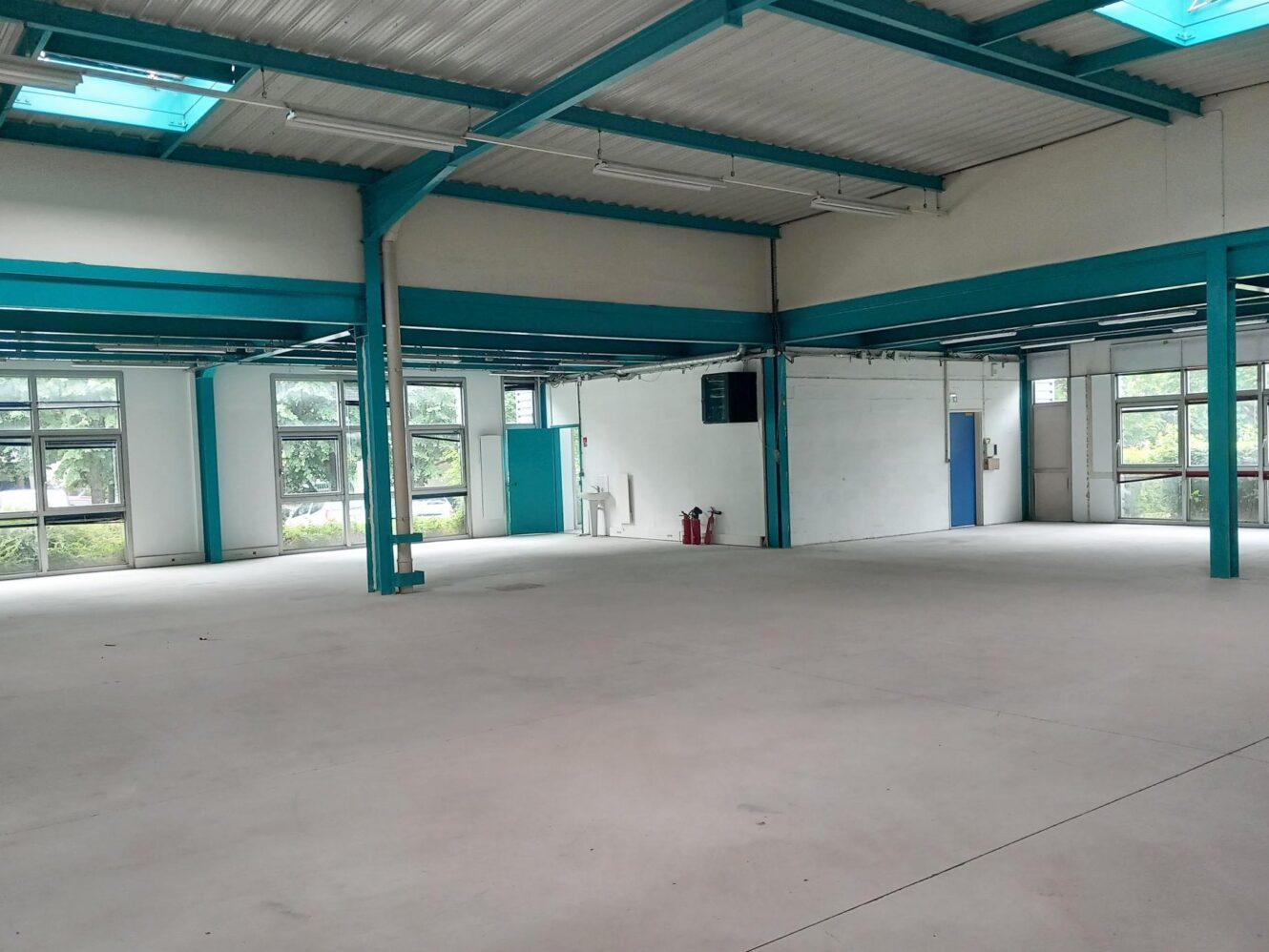 Intérieur industriel spacieux et vide, avec des poutres en acier bleu, de grandes fenêtres, des murs blancs et des installations minimales, notamment des extincteurs et un évier le long d'un mur.