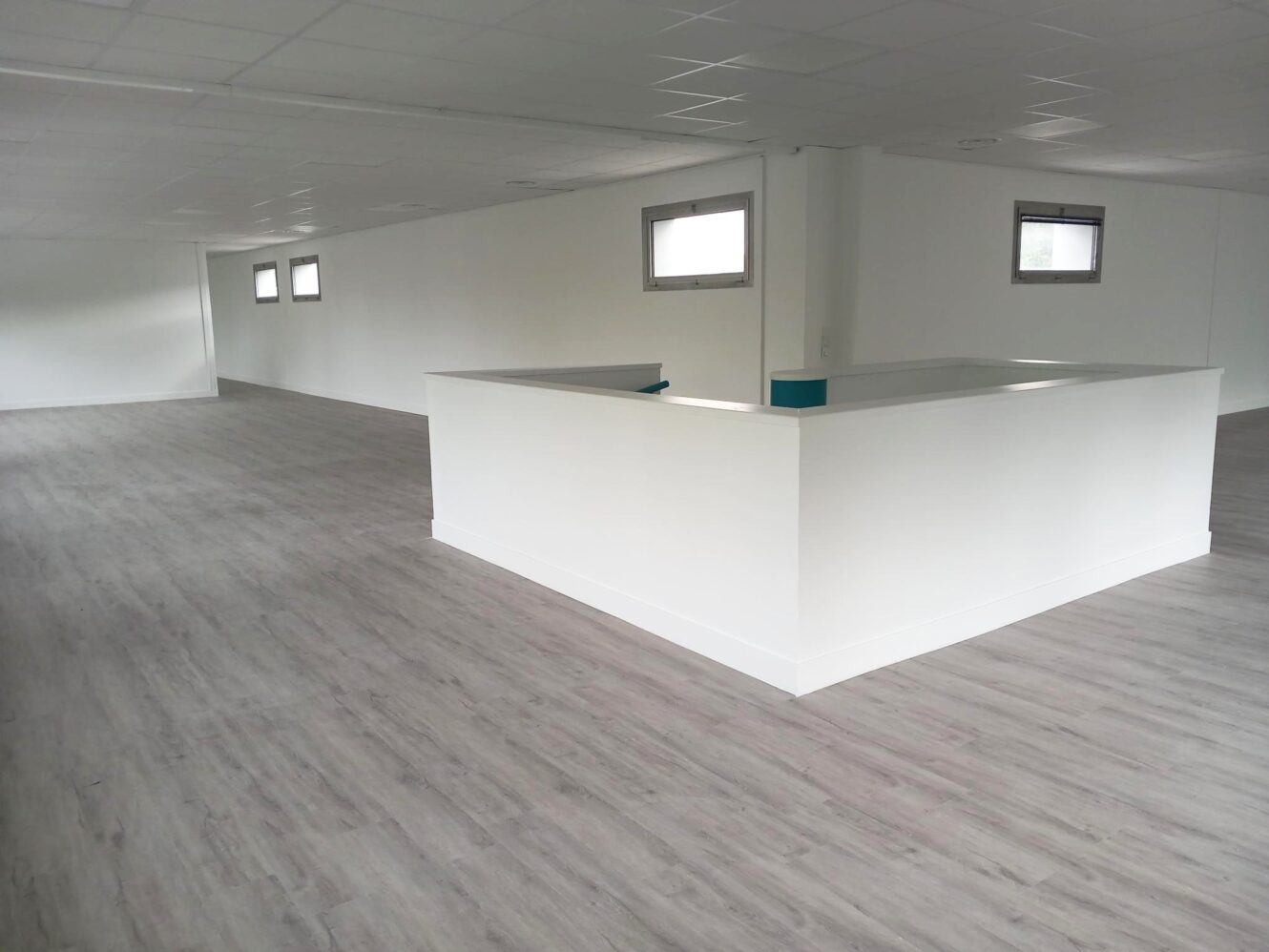 Espace de bureau vide avec un sol en bois gris clair, des murs blancs et une cloison centrale blanche sous de petites fenêtres rectangulaires.