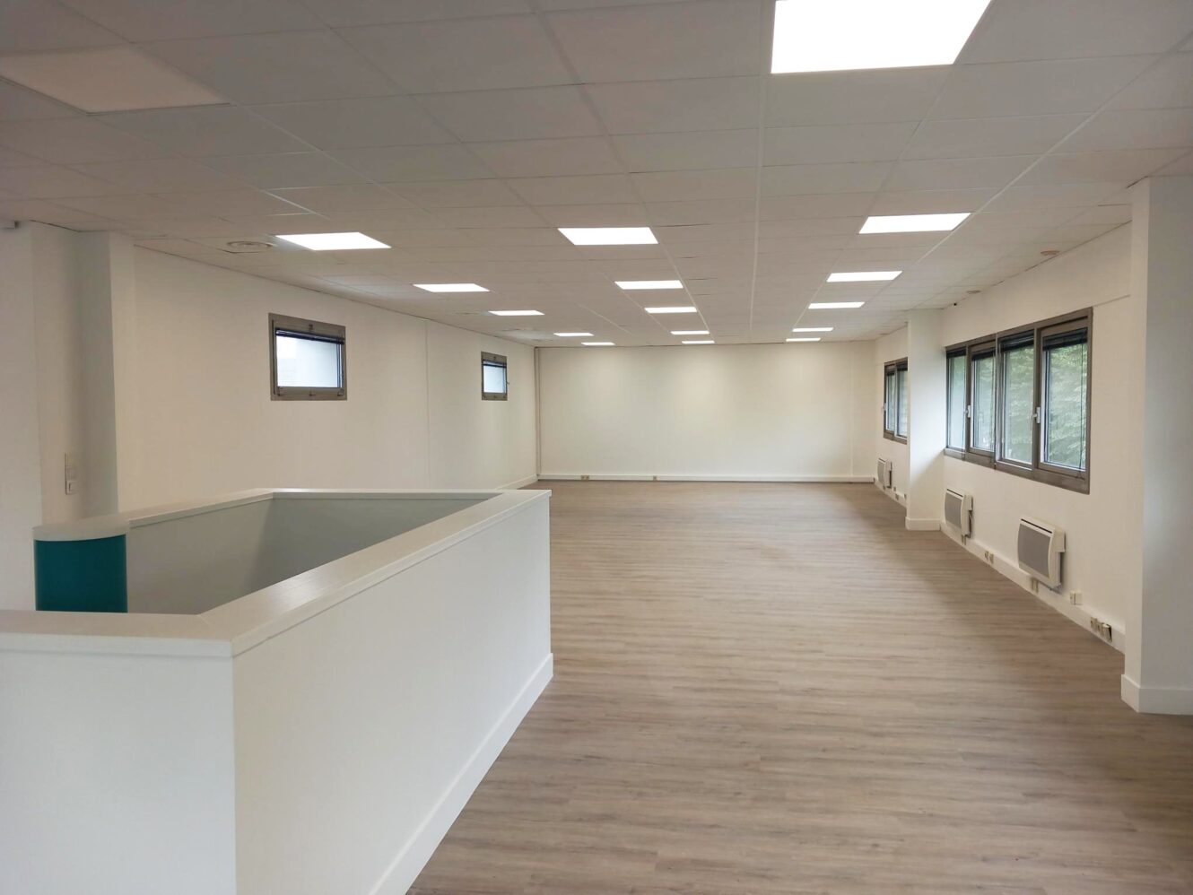 Espace de bureau vide et moderne avec des murs blancs, de grandes fenêtres, un parquet en bois clair et des plafonniers.