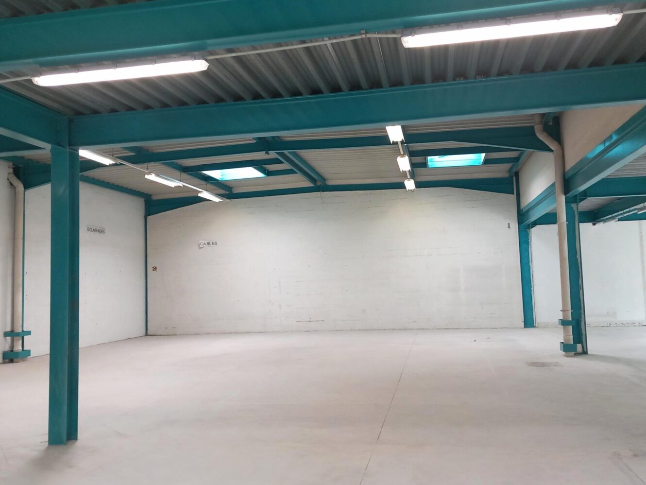Intérieur d'un entrepôt industriel vide avec des poutres en acier sarcelle, des murs blancs, un sol en béton et des plafonniers fluorescents.