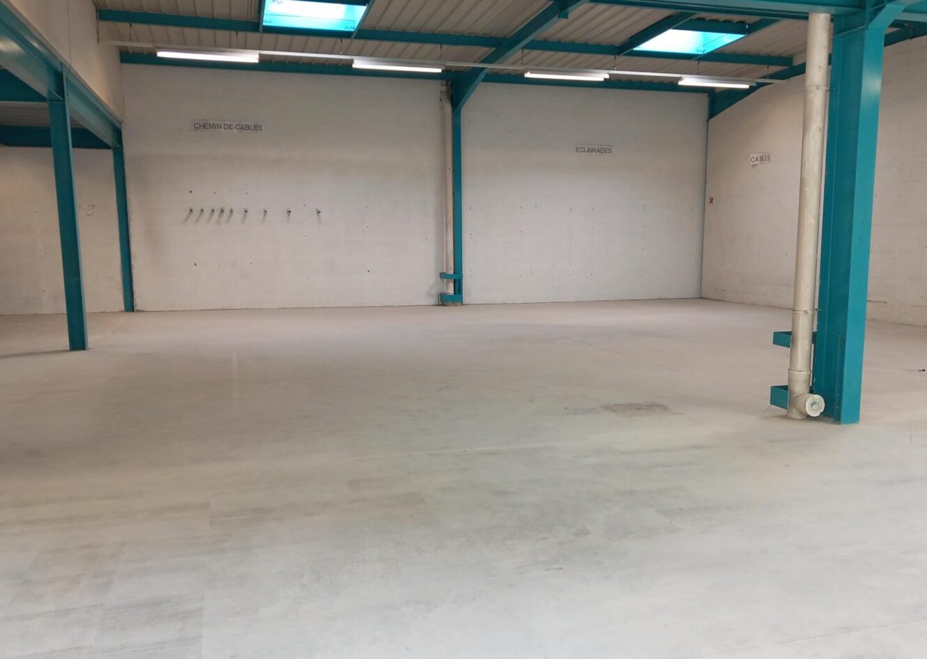 Entrepôt industriel vide avec des poutres de soutien sarcelles, des murs blancs et un sol en béton. Quelques crochets muraux et de petites enseignes sont visibles sur le mur du fond.