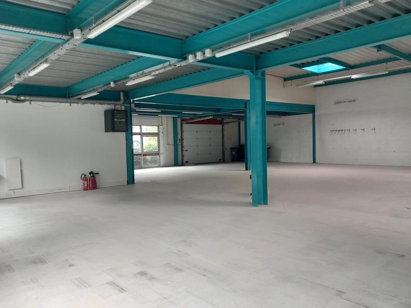 Intérieur d'un grand entrepôt industriel vide avec des poutres sarcelles, des sols en béton, des extincteurs et une grande porte de garage à l'arrière.