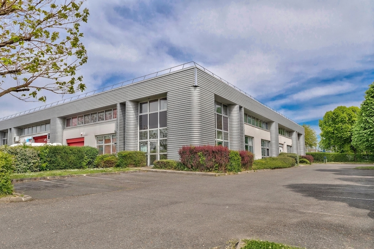 Un bâtiment commercial moderne, de plain-pied, doté de grandes baies vitrées et d'un bardage métallique, entouré d'arbustes et d'un parking vide, sous un ciel partiellement nuageux.