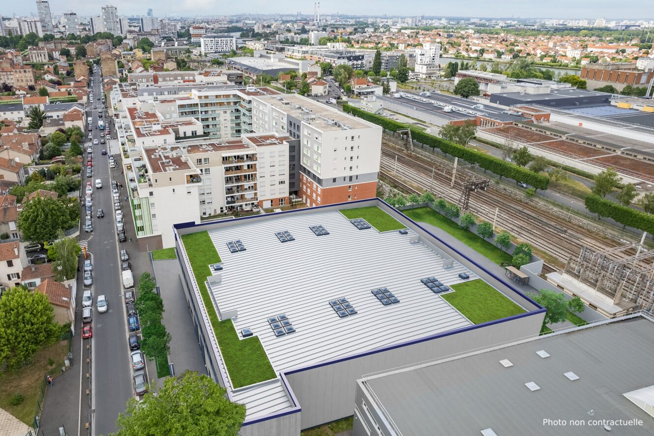 Vue aérienne d'un bâtiment industriel moderne avec un toit blanc et des espaces verts, entouré d'appartements résidentiels, de routes et de voies ferrées dans un environnement urbain.