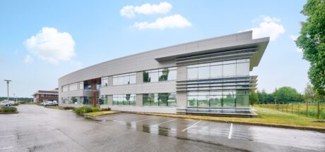 Immeuble de bureaux moderne avec de grandes baies vitrées, entouré de verdure et d'un parking humide vide, sous un ciel partiellement nuageux.