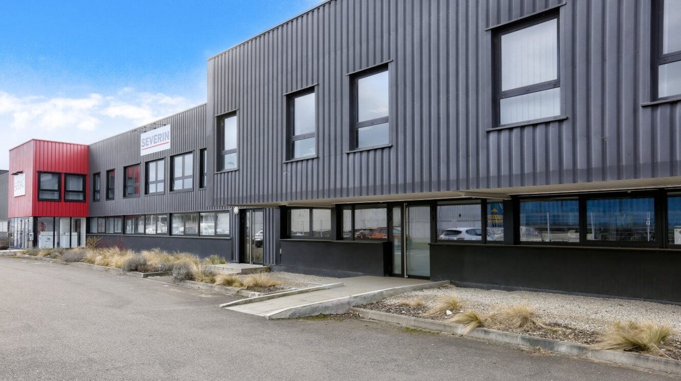 Bâtiment industriel ou de bureaux moderne avec des panneaux extérieurs noirs et rouges, plusieurs fenêtres et un petit espace paysager à l'avant.