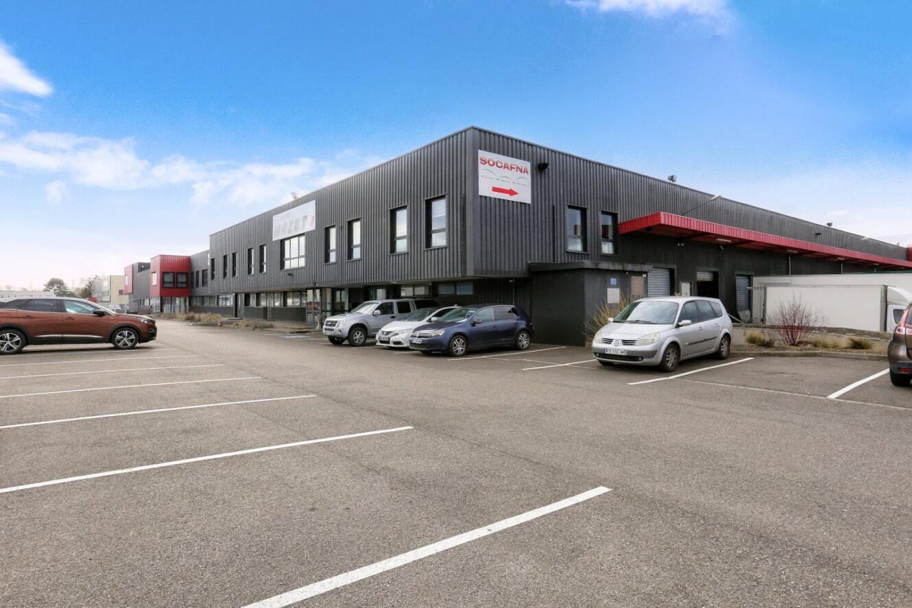 Un bâtiment industriel moderne avec un bardage gris foncé, plusieurs voitures garées et un parking en grande partie vide sous un ciel bleu.