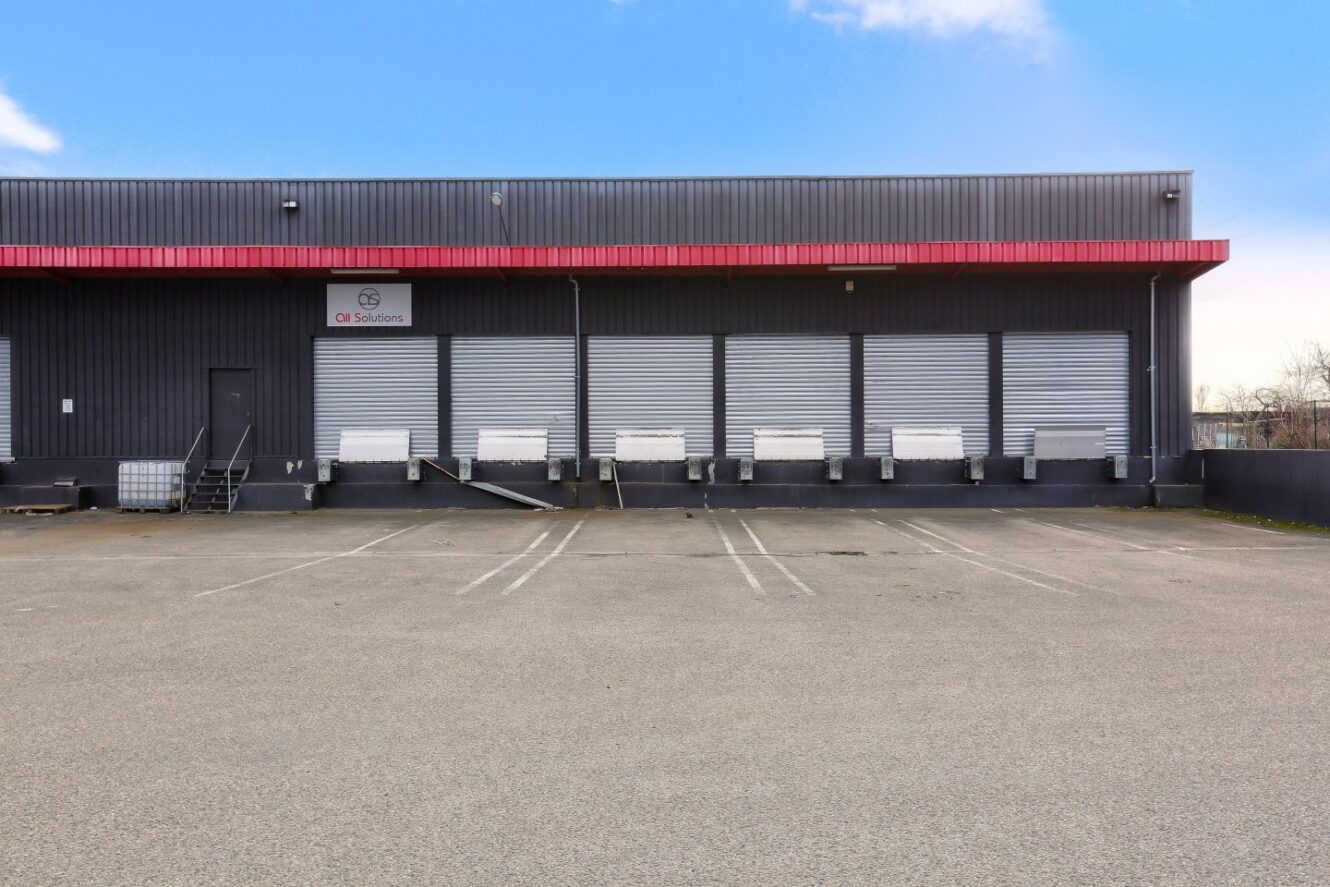 L'extérieur d'un entrepôt avec plusieurs portes de quai de chargement, un panneau indiquant CB Solutions et un parking vide devant l'entrepôt.