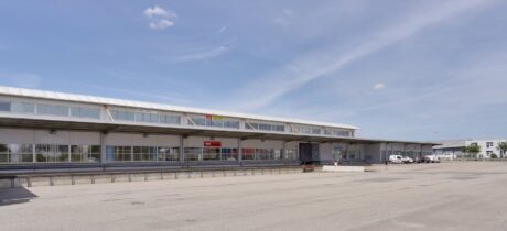 Un grand entrepôt industriel avec des quais de chargement, quelques véhicules garés et une zone pavée pratiquement vide devant, sous un ciel dégagé.