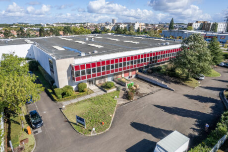 Vue aérienne d'un grand bâtiment industriel au toit plat et aux fenêtres rouges, entouré de voitures, d'arbres et de bâtiments urbains.