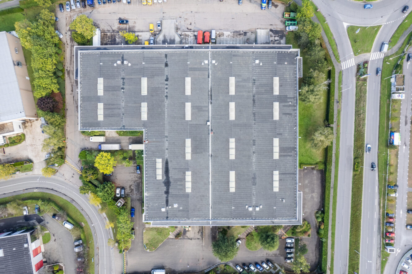 Vue aérienne d'un grand bâtiment industriel au toit gris, entouré de parkings, de voitures, de routes et de verdure.