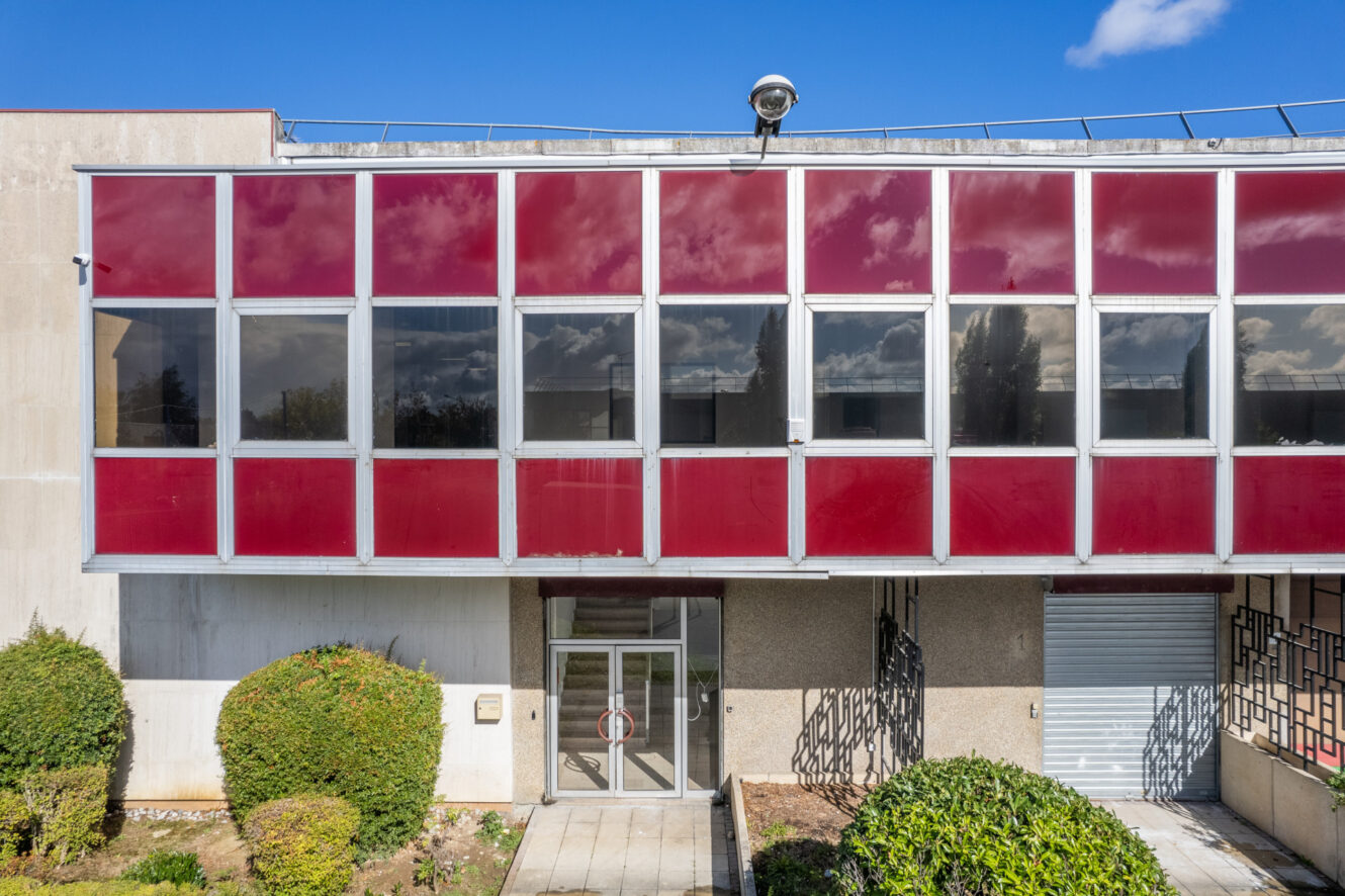 Une façade d'immeuble moderne avec de grandes fenêtres teintées de rouge, une porte d'entrée en verre, une porte de garage fermée et des buissons taillés devant sous un ciel bleu clair.