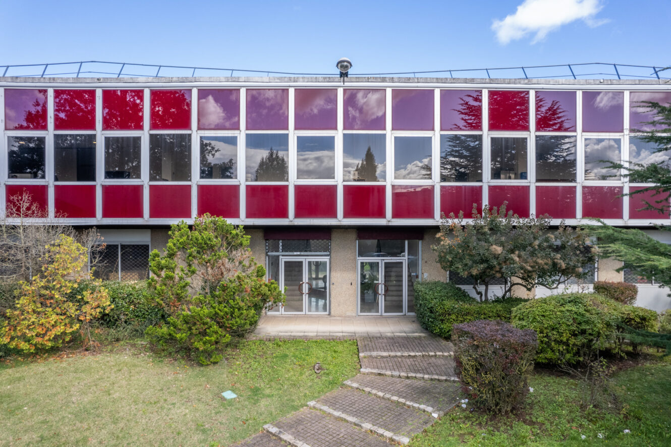 Un bâtiment de deux étages avec des fenêtres en verre rouge et réfléchissant, entouré d'arbustes verts et d'une pelouse avec une allée pavée menant à l'entrée.