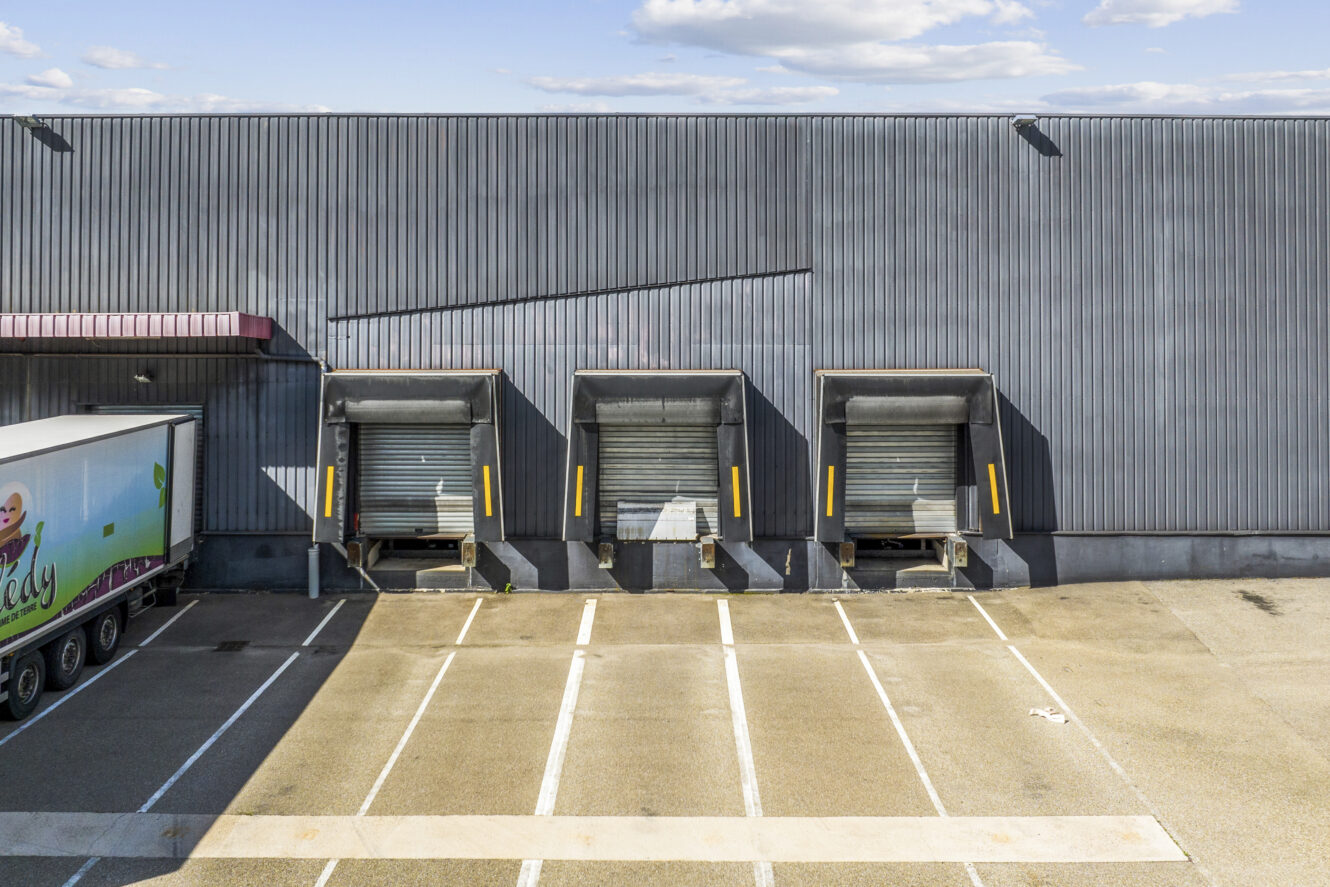 Trois quais de chargement avec des volets fermés à côté d'un parking vide ; un camion est partiellement visible sur le côté gauche du bâtiment industriel.