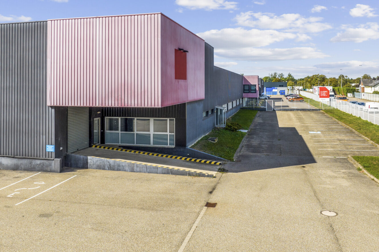 Bâtiment industriel avec extérieur rouge et gris, rampe de chargement, parking clôturé et ciel dégagé en arrière-plan.