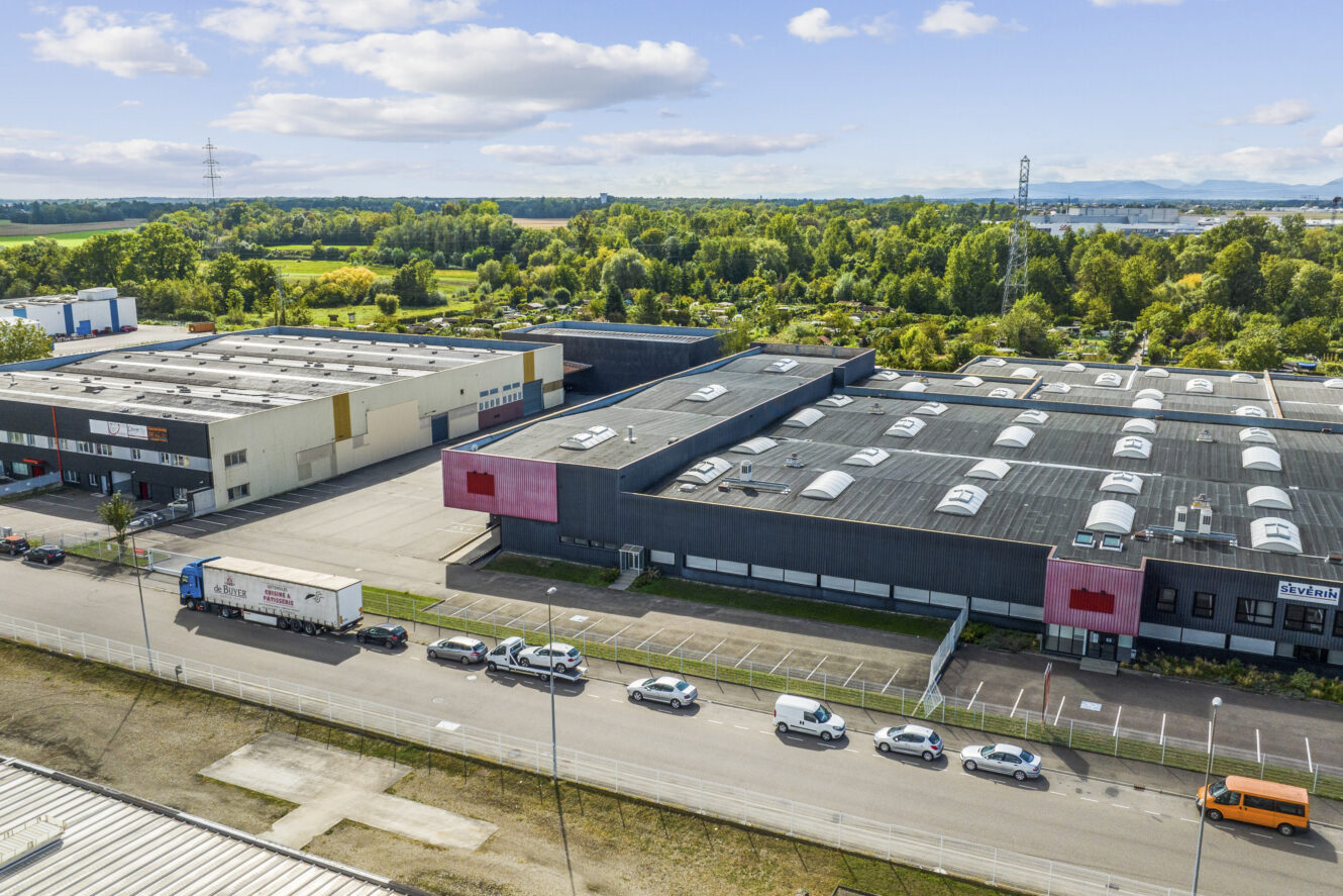 Vue aérienne d'une zone industrielle avec de grands entrepôts, des voitures garées le long de la rue, un camion et des arbres verts en arrière-plan, sous un ciel partiellement nuageux.
