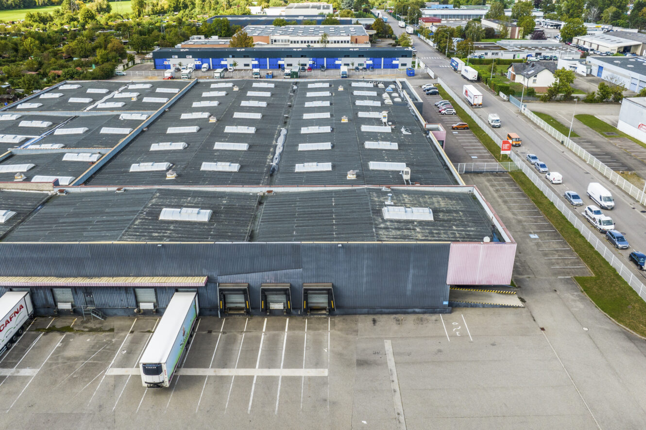 Vue aérienne d'un grand entrepôt industriel avec plusieurs quais de chargement, des camions en stationnement et des voitures dans les parkings adjacents et sur les routes environnantes.
