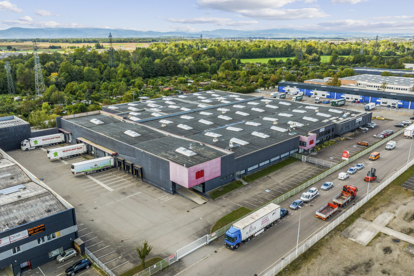 Vue aérienne d'un grand entrepôt industriel avec plusieurs quais de chargement, des camions et des voitures garées à l'extérieur, entouré de verdure et d'autres bâtiments industriels.
