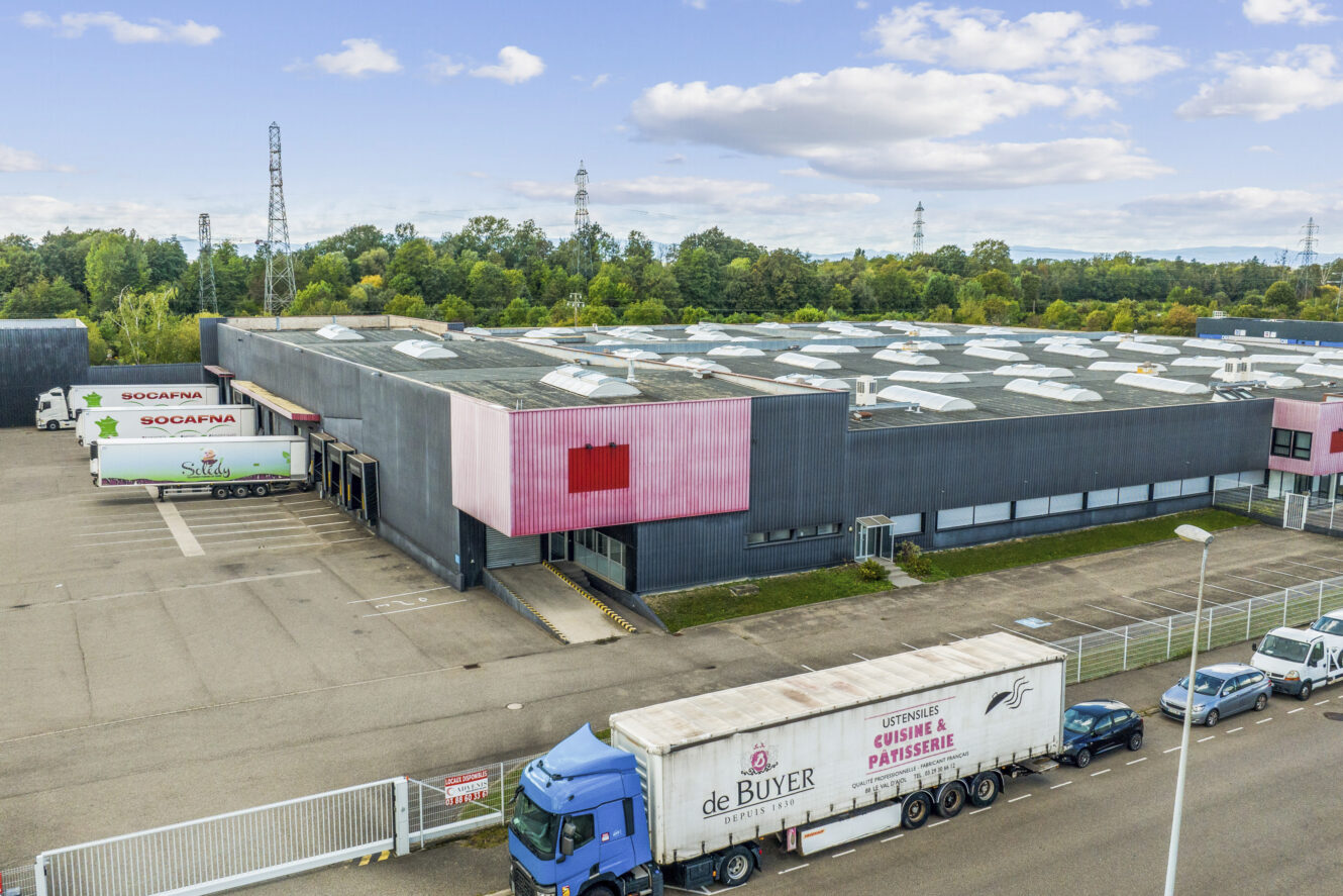 Entrepôt industriel avec des camions garés à l'extérieur, plusieurs quais de chargement, un périmètre clôturé et une zone forestière en arrière-plan, sous un ciel partiellement nuageux.