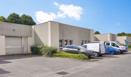 Bâtiment industriel de plain-pied avec une rangée de voitures et de camionnettes garées devant, par une journée ensoleillée avec un ciel bleu et des nuages épars.
