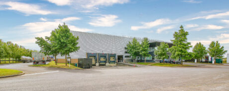 Un entrepôt industriel moderne avec des quais de chargement, des camions, des voitures garées et des arbres environnants sous un ciel partiellement nuageux.